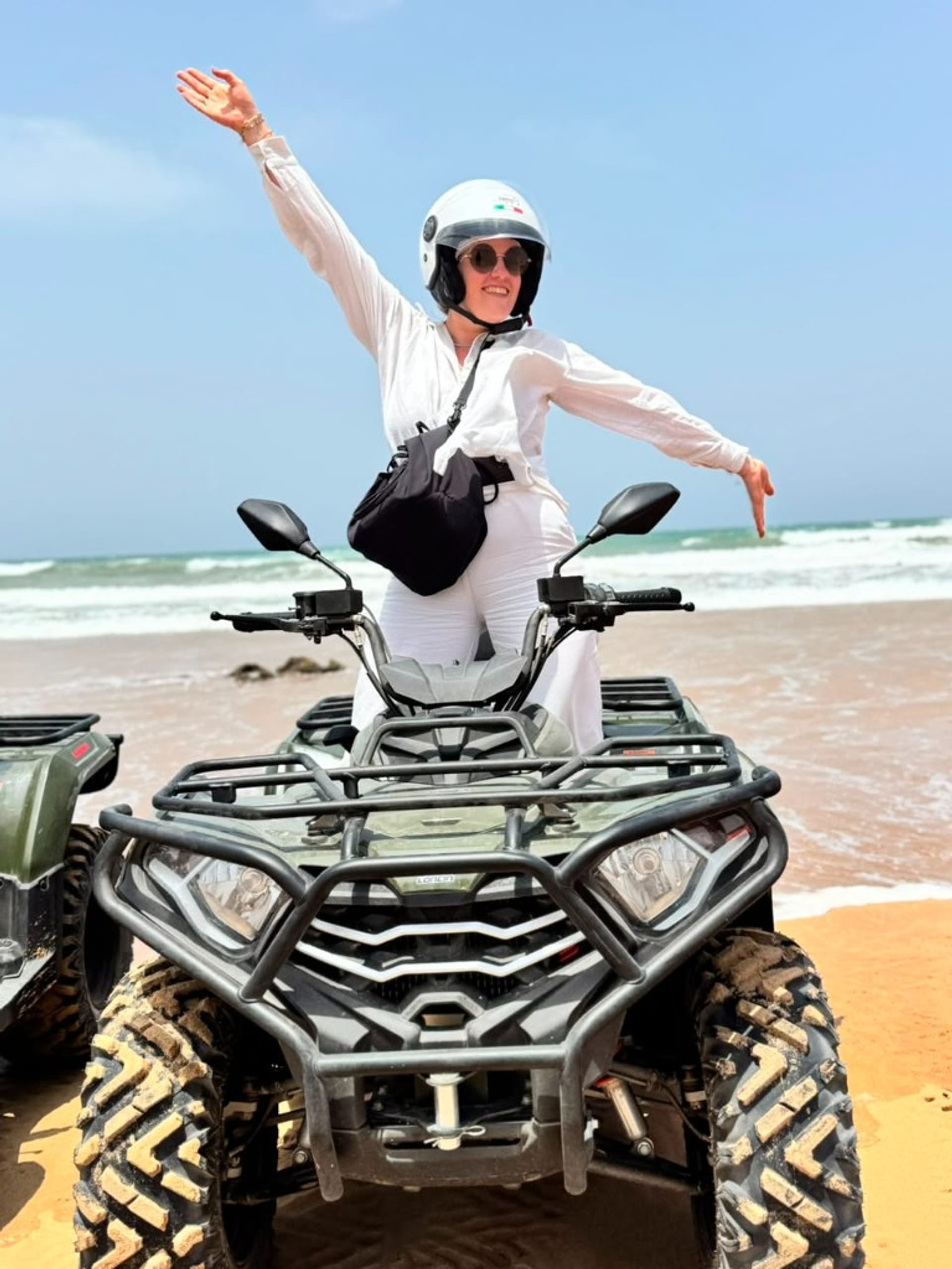 Femme en tenue blanche sur un quad, bras ouverts de joie sur la plage d'Essaouira face à l'océan